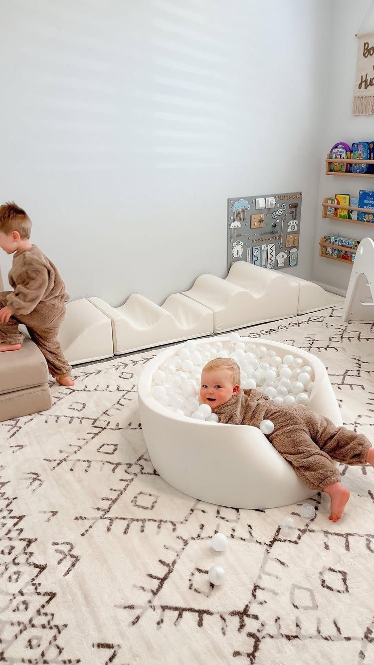 A toddler in brown pajamas relaxing inside a white ball pit filled with white balls on a patterned rug.