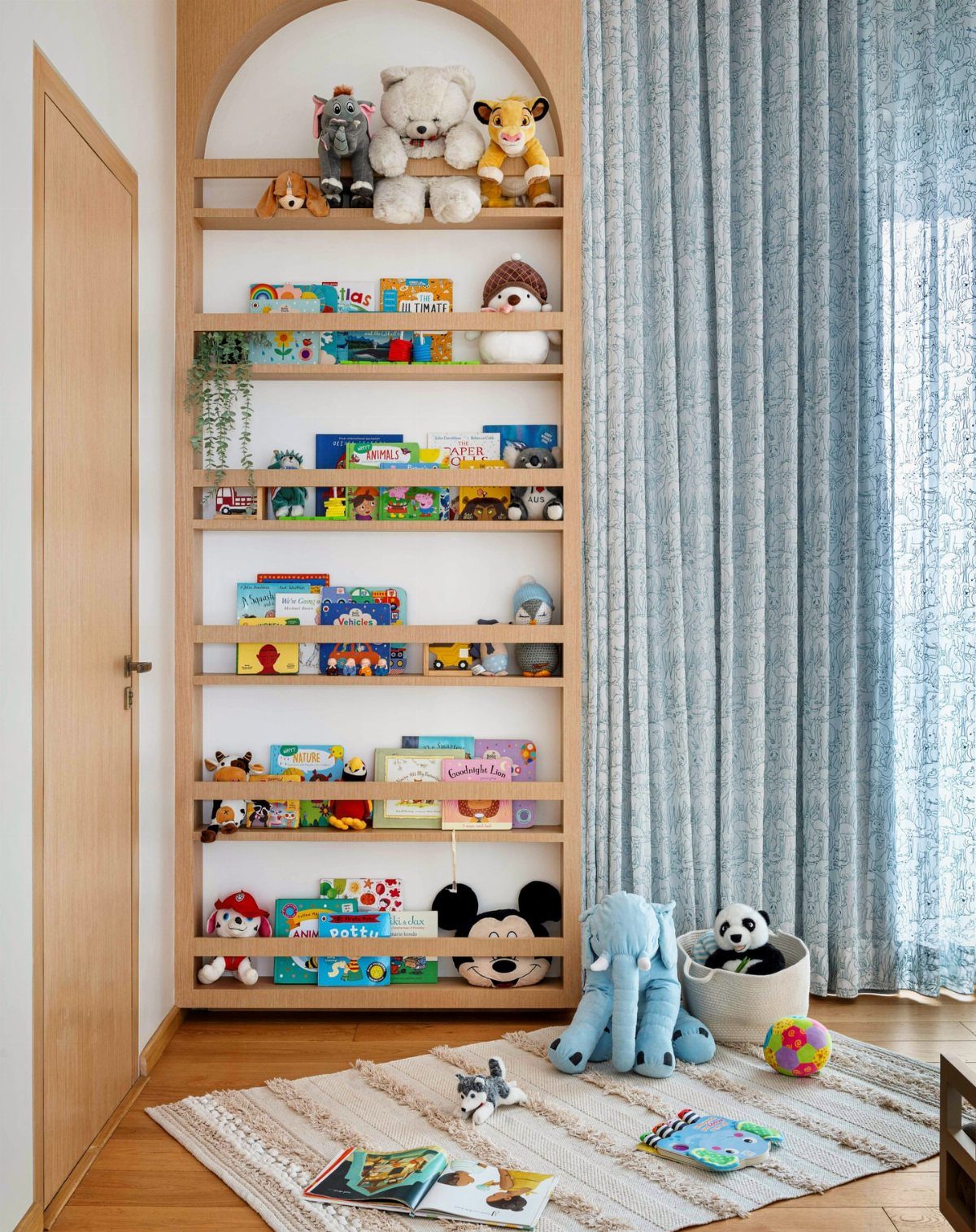 Wooden arched bookshelf displaying children's books and stuffed animals in a bright room.