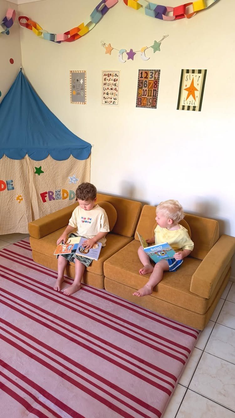 Two toddlers sitting on a yellow foam couch reading books in a playroom.