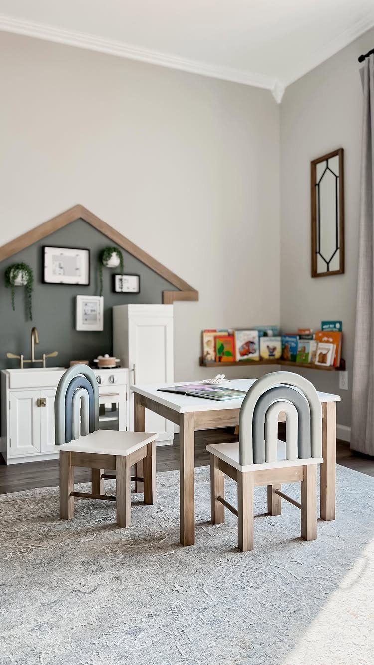 A white play kitchen, wooden table with rainbow chairs, and a bookshelf in a playroom.