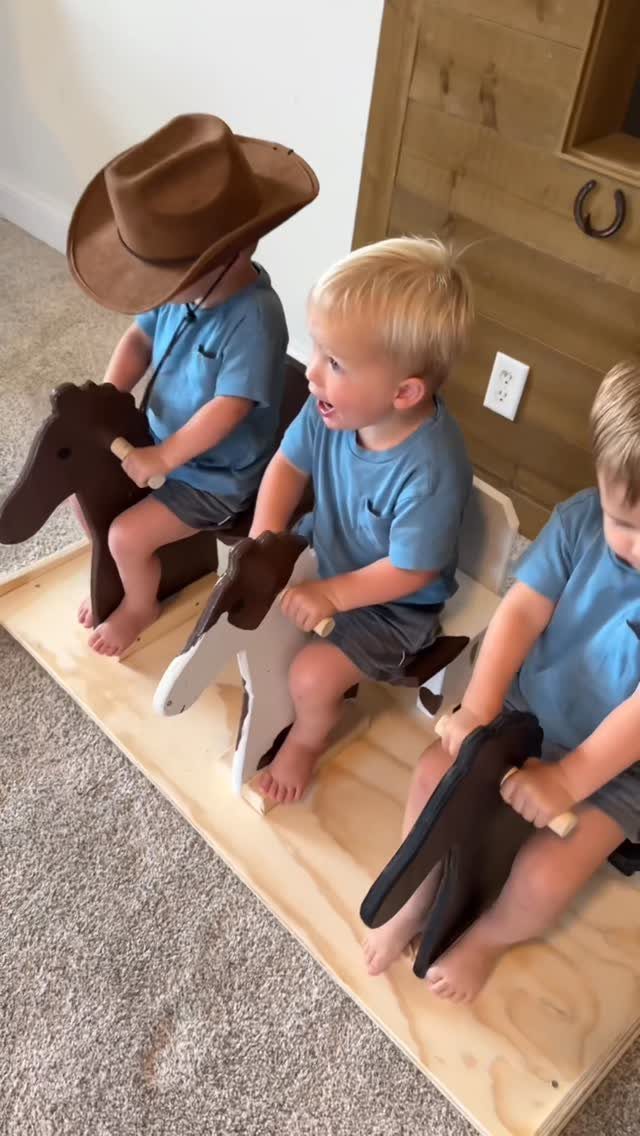 Three toddlers riding wooden rocking horses mounted on a single long wooden base.