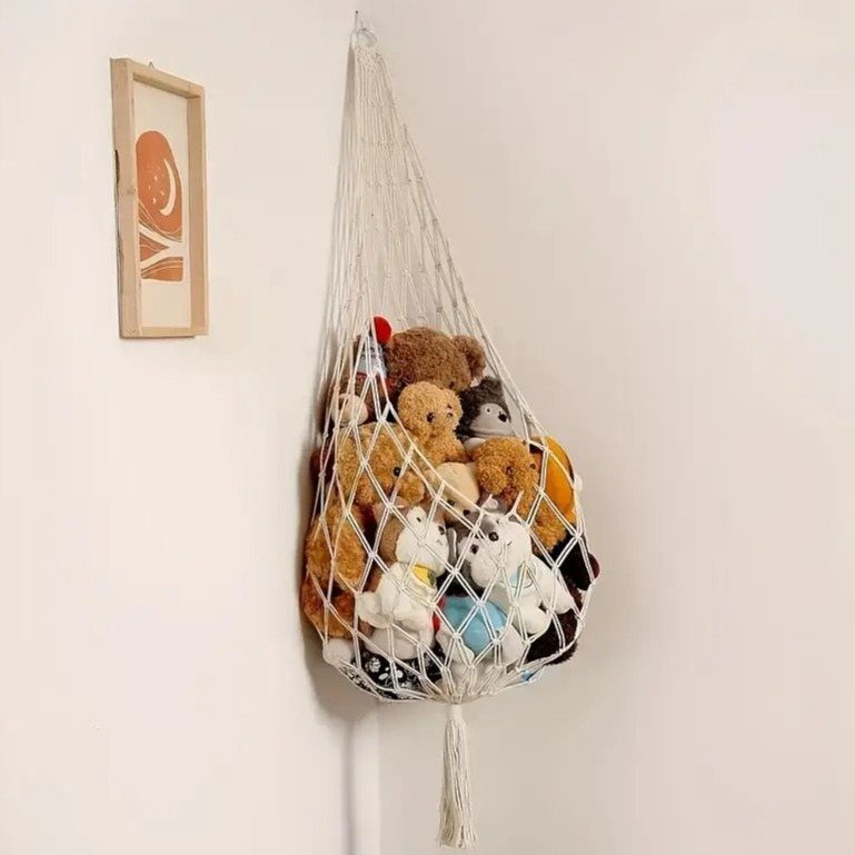 White macrame hanging net filled with various colorful stuffed animals mounted on a white wall.