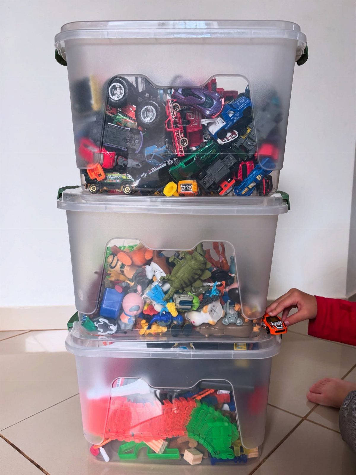 Three stacked clear plastic bins filled with colorful children's toys on a tiled floor.