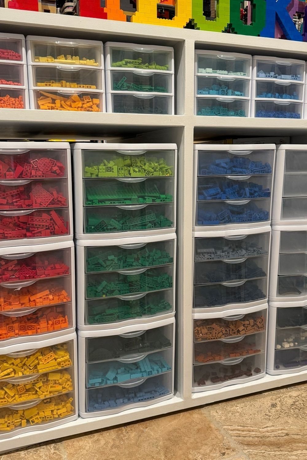Clear plastic drawers filled with color-sorted LEGO bricks inside a white storage shelving unit.