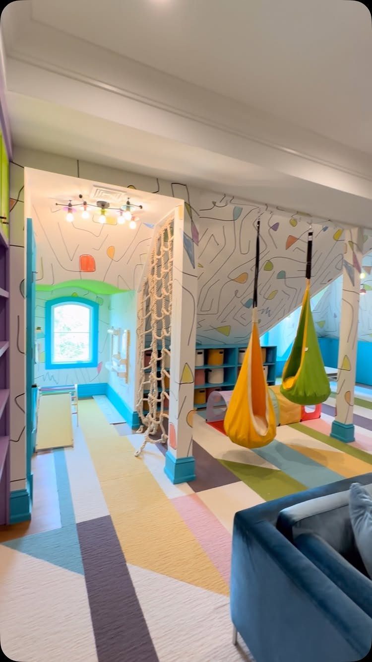 Colorful playroom with hanging cocoon swings, climbing nets, and geometric patterned rugs on the floor.