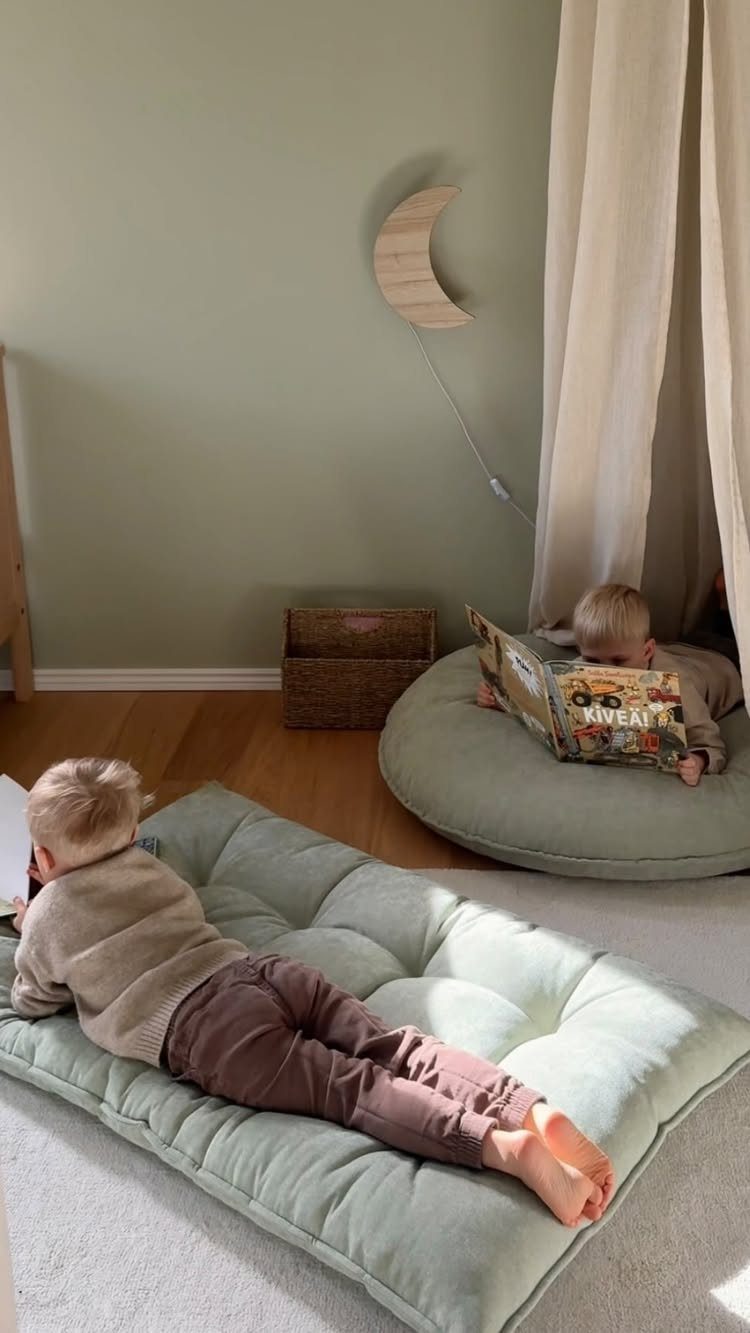 Two children lying on large, soft, sage green floor cushions while reading books.