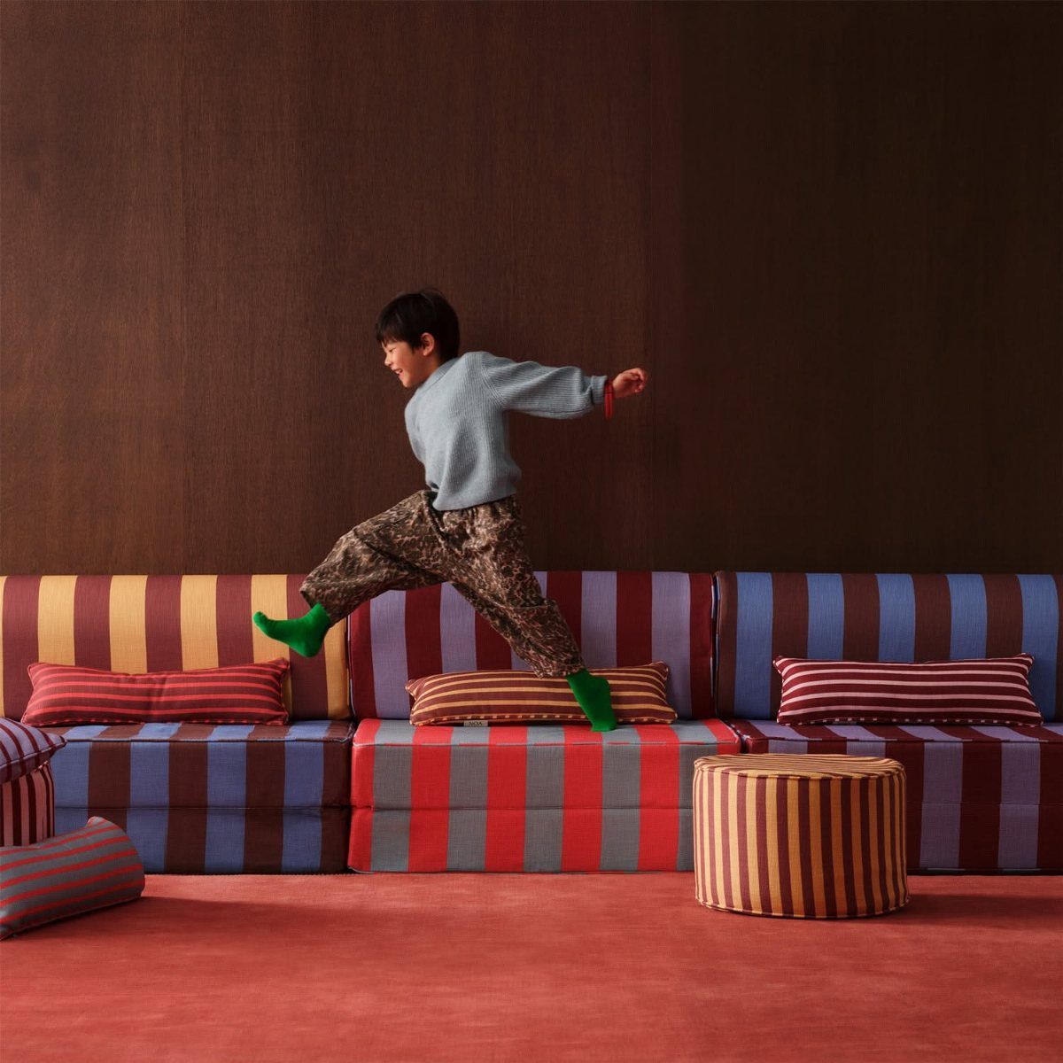 A child jumping over colorful striped modular sofa cushions in a room with red flooring.