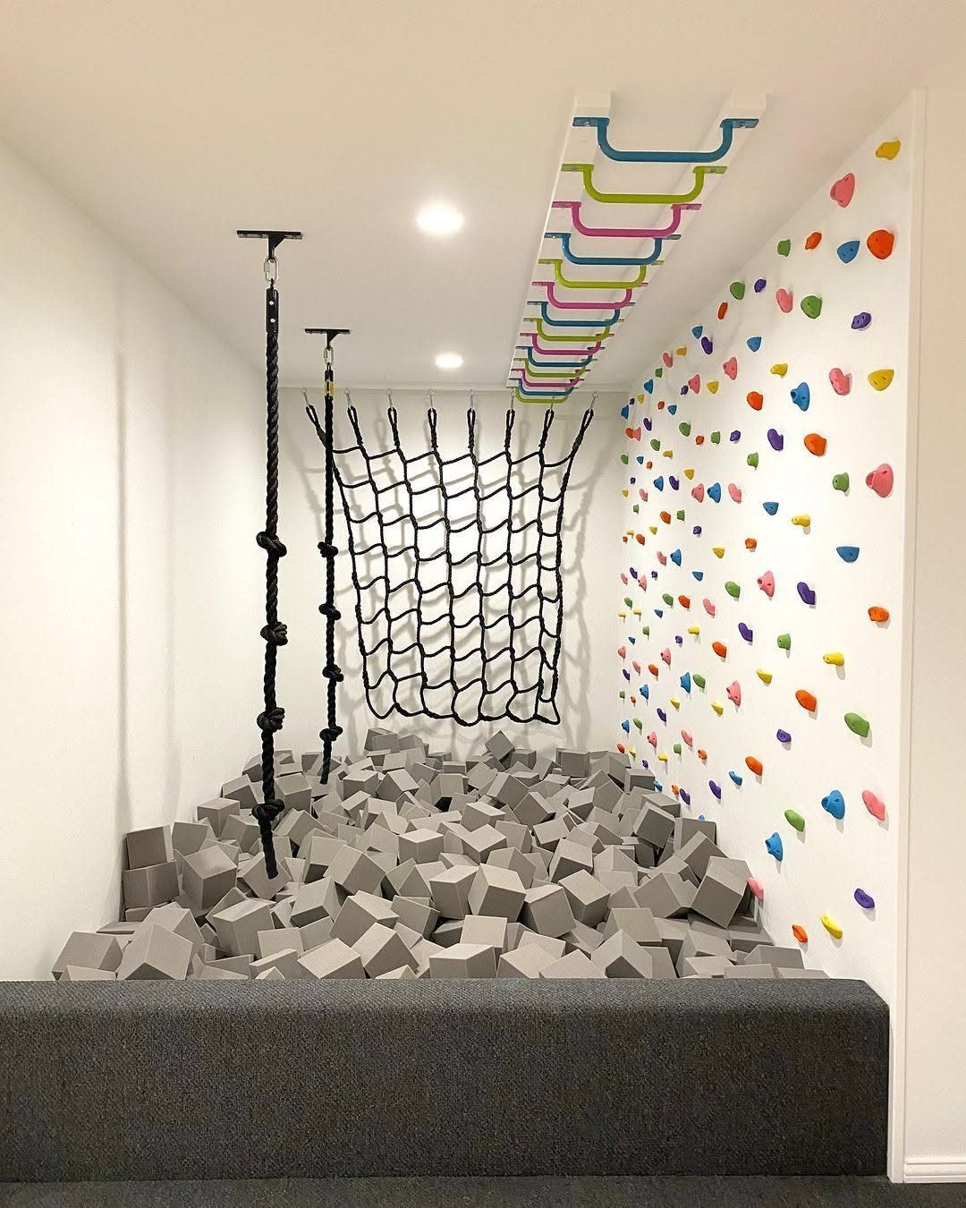 Indoor playroom with a colorful climbing wall, rope net, and grey foam cube pit.