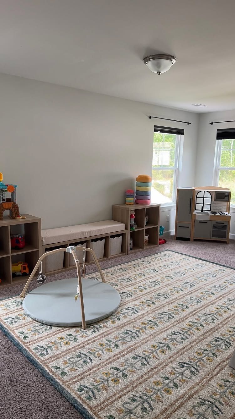 A playroom featuring wooden shelving units, storage bins, and a toy kitchen on a patterned rug.