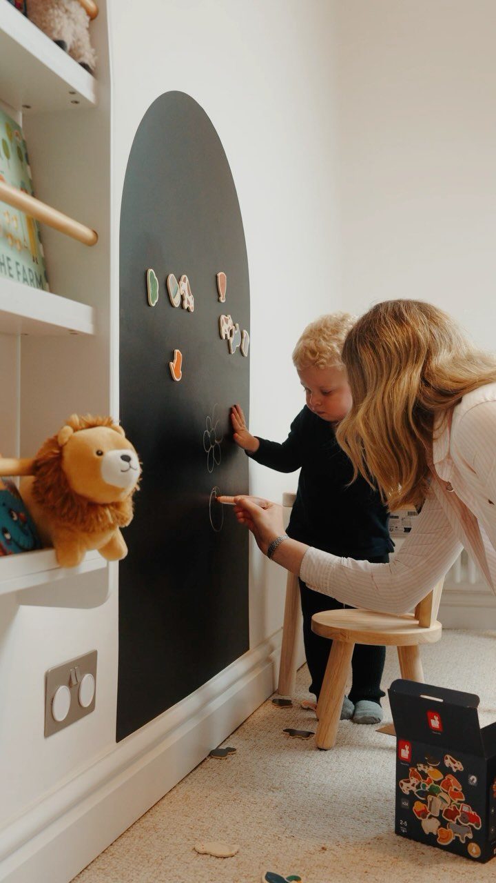 A child and woman drawing on a large black arch-shaped chalkboard wall in a playroom.