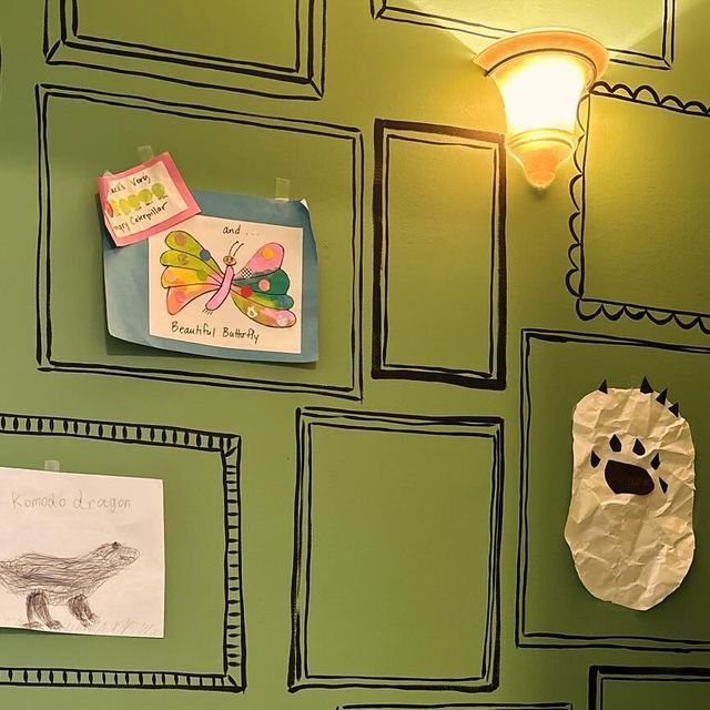 Children's drawings and paper crafts displayed inside hand-drawn black frames on a green wall.