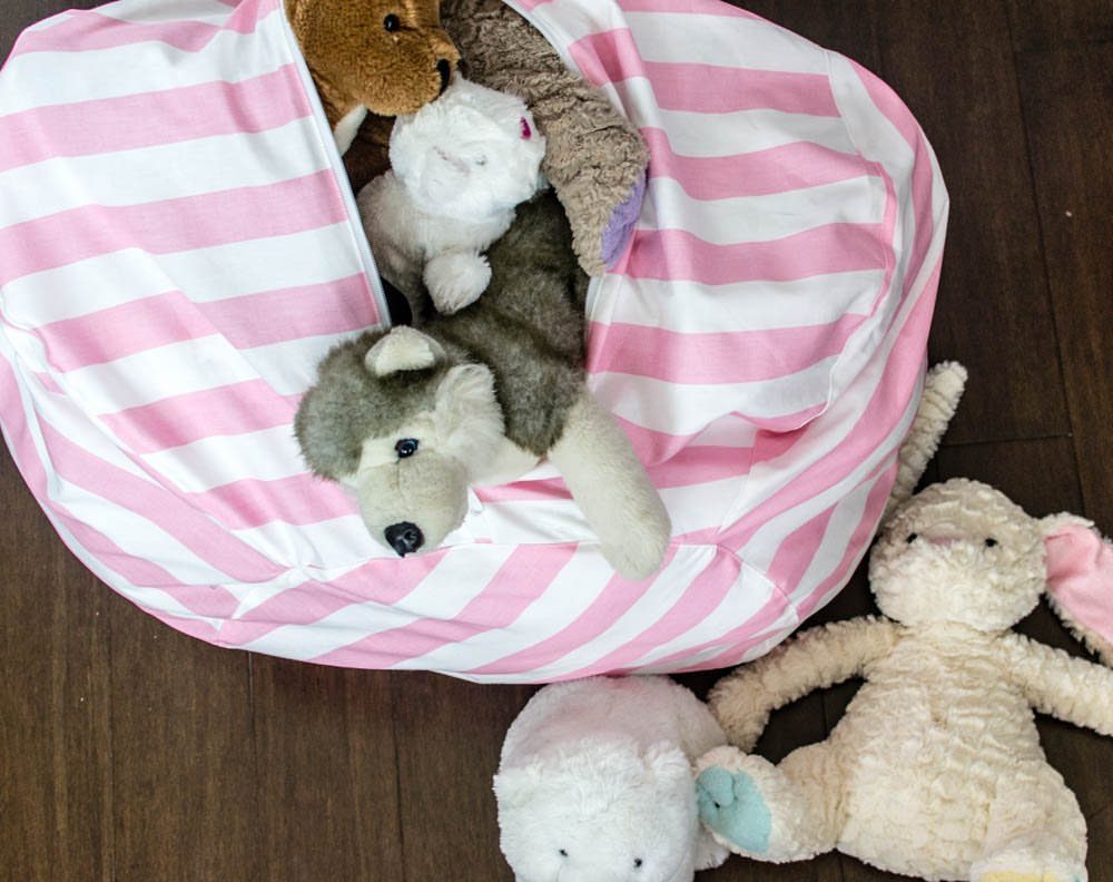 6 Plush Storage Ideas for Stuffed Toys That Clear Floor Pile-Ups 3 Pink and white striped bean bag overflowing with stuffed animals on a dark wood floor.