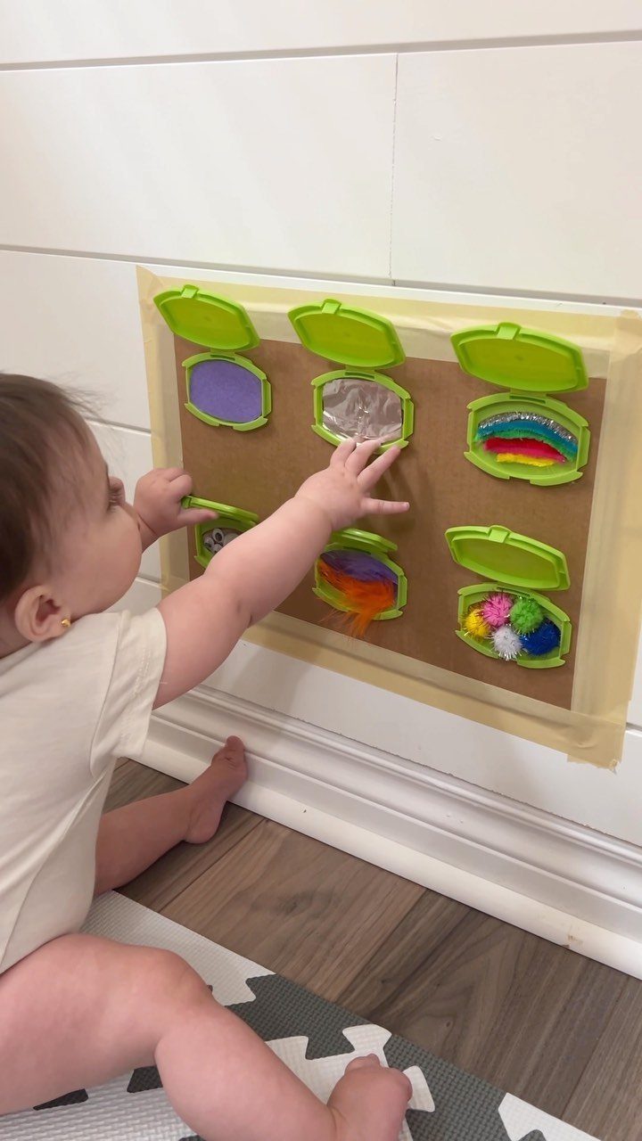 23 Small Playroom Design Ideas That Fit Real Rooms 7 A baby reaches toward a wall-mounted sensory board with various colorful textures and materials.