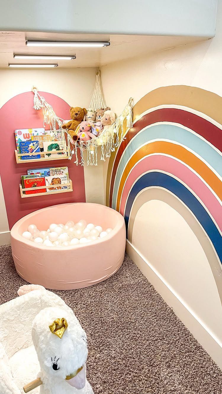 23 Small Playroom Design Ideas That Fit Real Rooms 1 A pink ball pit sits below wall-mounted bookshelves and a macrame toy hammock.