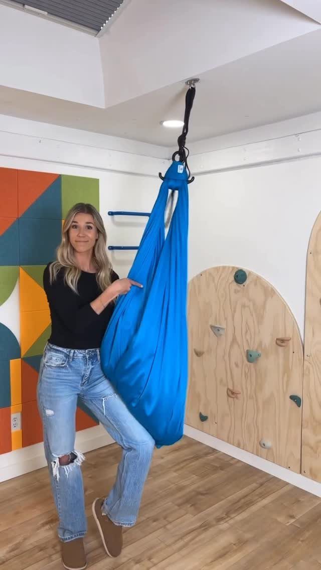 A woman pointing to a bright blue fabric swing hanging from the ceiling in a playroom.