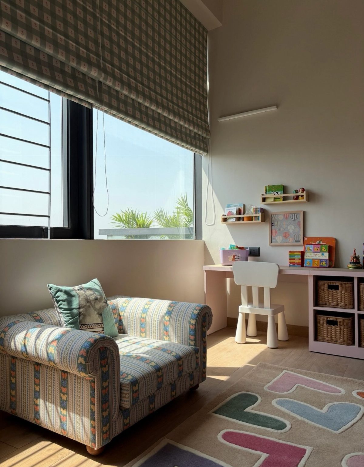 A bright playroom with a large window, patterned armchair, white desk, and colorful rug.