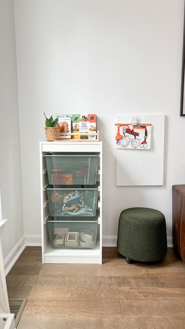 White storage unit with three mesh baskets containing toys, books, and a green ottoman on a wooden floor.