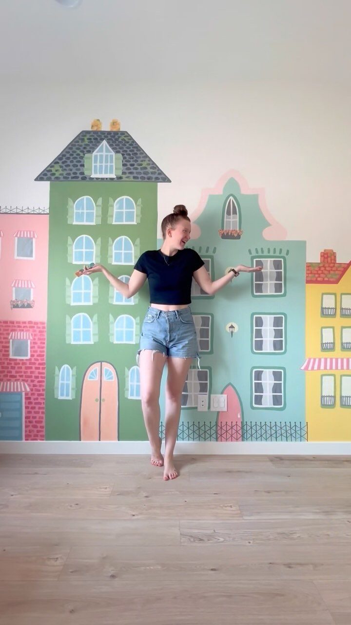 A woman posing in front of a colorful townhouse wall mural in a room.