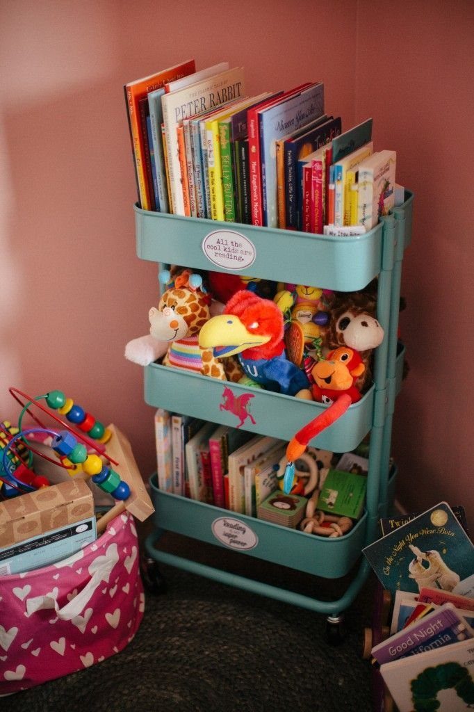 7 Toy Organization Ideas for Playrooms and Living Spaces 4 A teal three-tier rolling cart filled with colorful books and stuffed animals in a pink room.